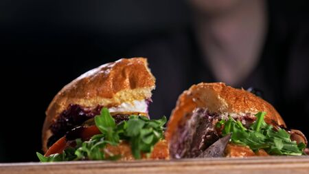 Cut craft burger is cooking on black background. Consist: sauce, arugula, tomato, red onion, bacon, red currant sauce, ricotta cottage fresh cheese, air bun and marble meat beef. Not made ideal. Looks real, loving hand made.の写真素材