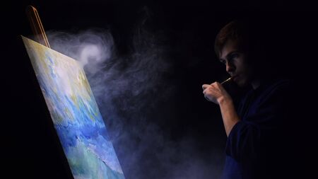 Artist copyist paint seascape with ship in ocean. Vaper smoke vape e-cigarette. Craftsman decorator draw as boat sail on blue sea with acrylic oil color. Draw finger, brush, knife palette. Indoor.の写真素材