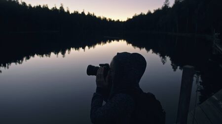 Silhouette traveler photographing scenic view in forest, river. Wood pier. One woman shooting nice dark magic night look. Girl take photo video on camera. Photographer walk with backpack. Outdoor.の写真素材