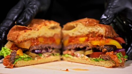 Cut craft burger is cooking on black background in black food gloves. Consist: red sauce salsa, lettuce, red onion, pickle, cheese, chilli green pepper, air bun and marbled meat beef. Not made ideal. Looks real, loving hand made.の写真素材