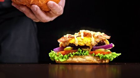 Craft burger is cooking on black background. Consist: sauce, lettuce, tomato, red onion, pickle, cheese, bacon, air bun and marbled meat beef. Not made ideal. Looks real, loving hand made.の写真素材