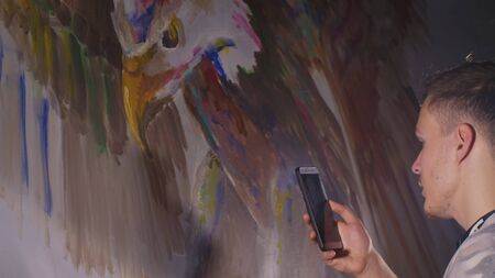 Artist designer draws an eagle on wall. Craftsman decorator paints picture with acrylic oil color looking at sketch in phone. Painter painter dressed in paint coat. Indoor. Dark magic cinematic look.の写真素材