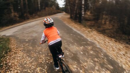 One caucasian children rides bike road in autumn park. Little girl riding black orange cycle in forest. Kid goes do bicycle sports. Biker motion ride with backpack and helmet. Mountain bike hardtail.の写真素材