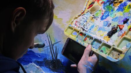 Artist copyist paint seascape with ship in ocean. Craftsman decorator draw as boat sail on blue sea with acrylic oil color. Draw finger, brush, knife palette. In hand of cellphone tablet computer pc.の写真素材