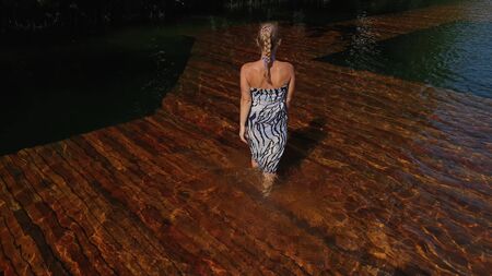 Woman walk on water on pier in sunglasses and a boho silk shawl. Girl rest on a flood wood underwater dock. The pavement is covered with water in the lake. In the background are mountain and a forest.の写真素材
