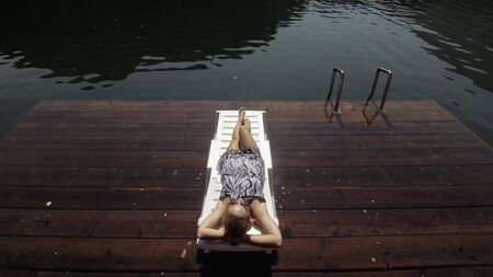 Woman lie on a sunbed in sunglasses and a boho silk shawl. Girl rest on a flood wood underwater pier. The pavement is covered with water in the lake. In the background are mountain and a forest.の写真素材