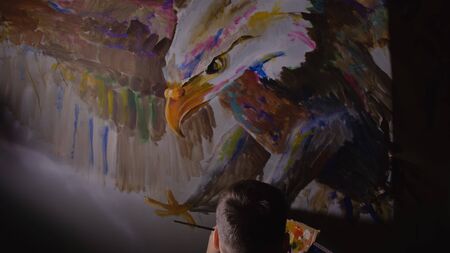 Artist designer draws an eagle on wall. Craftsman decorator paints picture with acrylic oil color looking at sketch in phone. Painter painter dressed in paint coat. Indoor. Dark magic cinematic look.の写真素材