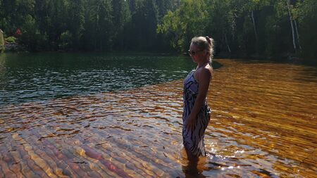 Woman walk on water on pier in sunglasses and a boho silk shawl. Girl rest on a flood wood underwater dock. The pavement is covered with water in the lake. In the background are mountain and a forest.の写真素材