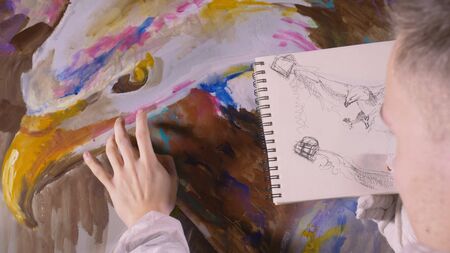 Artist designer draws an eagle on wall. Craftsman decorator paints picture with acrylic oil color looking at sketch in phone. Painter painter dressed in paint coat. Indoor. Dark magic cinematic look.の写真素材