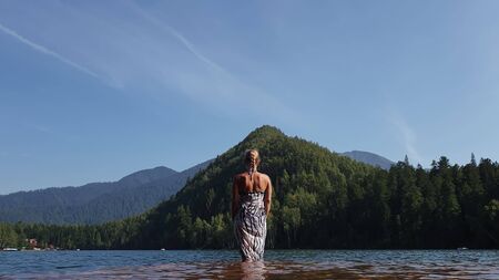 Woman walk on water on pier in sunglasses and a boho silk shawl. Girl rest on a flood wood underwater dock. The pavement is covered with water in the lake. In the background are mountain and a forest.の写真素材