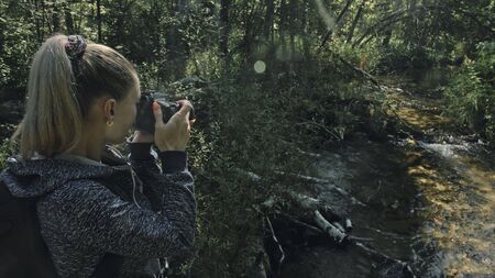 Traveler photographing scenic view in forest river. Wood bridge fallen tree. One caucasian woman shooting nice magic look. Girl take photo video on camera. Photographer travel with backpack. Outdoor.の写真素材
