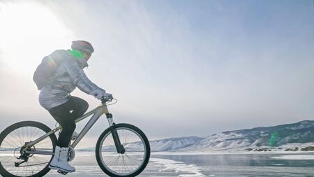 Woman is riding bicycle on the ice. Girl is dressed in a silvery down jacket, cycling backpack and helmet. Ice of the frozen Lake Baikal. Tires on bike are covered with spikes. Traveler is ride cycle.の写真素材
