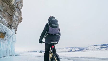 Man is riding bicycle near ice grotto. Rock with ice caves icicles. Teenage is dressed in black down...