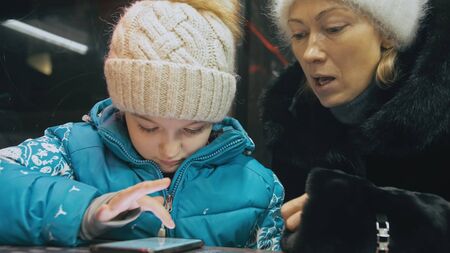 Attractive caucasian mother and daughter using smartphone sitting in winter cafe in jacket, hat. Advanced little child girl shows and explains to mom woman on smartphone, touching and watching.の写真素材