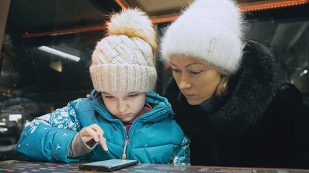 Attractive caucasian mother and daughter using smartphone sitting in winter cafe in jacket, hat. Advanced little child girl shows and explains to mom woman on smartphone, touching and watching.の写真素材