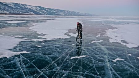 Man is riding bicycle on the ice. Ice of the frozen Lake Baikal. Teenage is dressed in black down jacket, cycling backpack, helmet. Tires on covered with special spikes. Traveler boy is ride cycle.の写真素材
