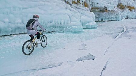 Woman is riding bicycle near the ice grotto. The rock with ice caves and icicles. Girl is dressed in silvery down jacket, cycling backpack and helmet. Tires winter with spikes. Traveler is ride cycle.の写真素材