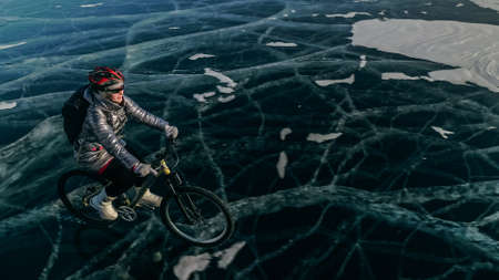Woman is riding bicycle on the ice. Girl is dressed in a silvery down jacket, cycling backpack and helmet. Ice of the frozen Lake Baikal. Tires on bike are covered with spikes. Traveler is ride cycle.の写真素材