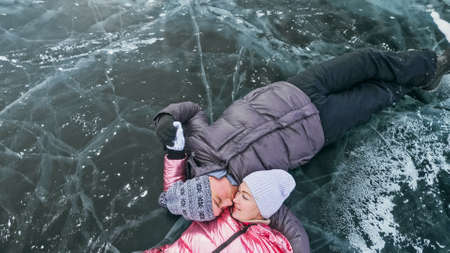 Couple has fun winter walk against background of ice of frozen lake. Lovers lie on clear ice with cracks have fun kiss and hug. View from above. Happy people on snow covered ice. Honeymoon love story.の写真素材