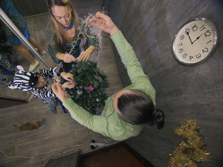 The family fast and fanny decorates the Christmas tree. Top view. Time on the clock is 23.45 on wall. Happy dad, mom and daughter are running around tree, hanging colored balls, garlands.の写真素材