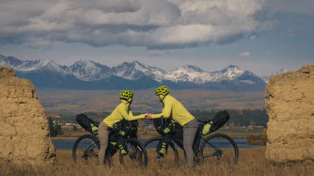 The man and woman travel on mixed terrain cycle bike touring with bikepacking. The two people journey with bicycle bags. Sport sportswear in green black colors. Mountain snow capped, stone arch.の写真素材