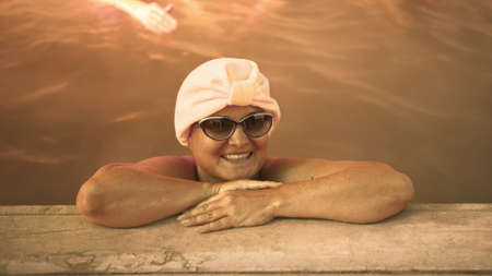 Close up caucasian woman in a swim in hot saline mineral water bath at a traditional spa outdoor with soft magic pink lighting and steam. Girl relaxing spring in hot geothermal spa pool is towel cap.の写真素材