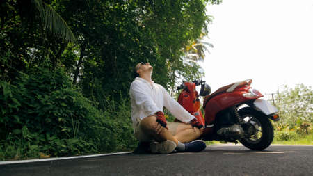 Man on red motorbike in white clothes drive on forest road trail trip. One men caucasian tourist in sunglasses sit on pavement, relax, rest, near scooter. Asia Thailand ride tourism. Motorcycle rent.の写真素材