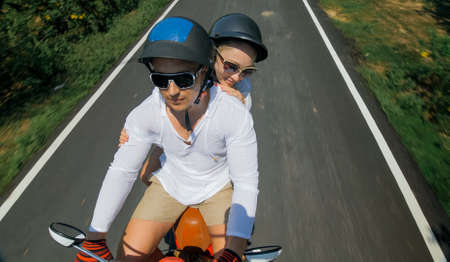 Two caucasian tourist woman man drive on red scooter. Make it selfie. Love couple on motorbike in white clothes to go on forest road trail trip. Motorcycle rent safety helmet sunglasses. Ride tourism.の写真素材