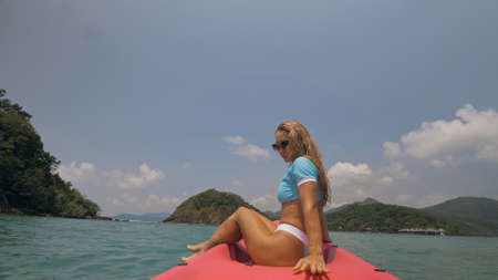Pretty young blonde woman in stylish swimsuit and sunglasses sits on pink canoe sailing along sea...