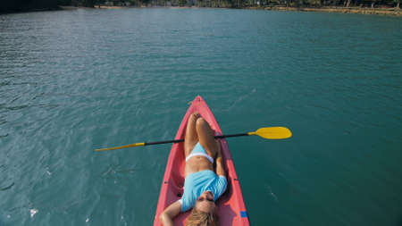 Pretty slim woman in elegant swimsuit lies in pink plastic kayak drifting on azure ocean at exotic...
