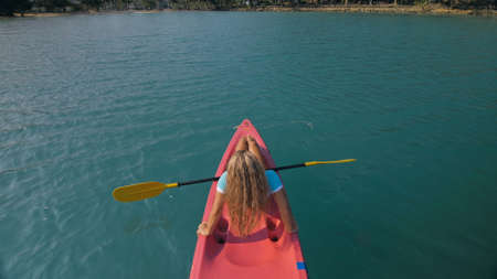 Seductive blonde woman in blue swimsuit sails on pink plastic kayak along azure sea at exotic resort...