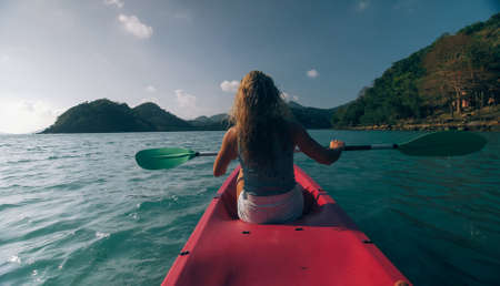 Silhouette of woman tourist raising hands while sailing in canoe along sea bay water to distant hilly island backside view.の写真素材