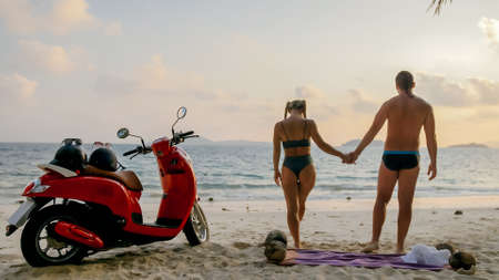 Scooter road trip. Lovely couple on red motorbike in white clothes on sand beach. People walking near the tropical palm trees, sea. Motorcycle rent.の写真素材