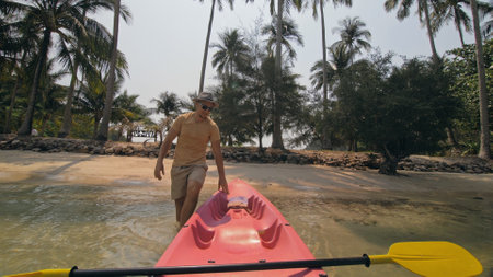 Man with sunglasses and hat rows pink plastic canoe along sea against green hilly islands with wild jungles. Traveling to tropical countries.の写真素材