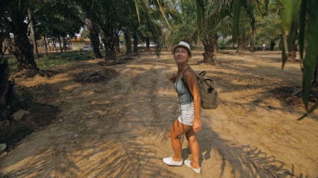 Woman tourist with plait walks looking around at growing young trees with lush leaves at oil palm...