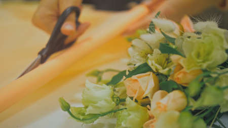 Female florist makes flower bunch, she cuts off wrapping paper working in flower shop. Cute young...