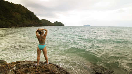 Woman chill relax resting on rock of sea reef stone, stormy cloudy ocean. Woman in swimsuit dress tunic. Concept rest tropical outdoor tourismの写真素材