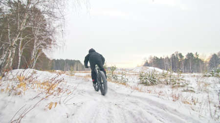 Professional extreme sportsman biker riding fat bike in outdoors. Cyclist ride in winter in snow field, forest. Man does trick on mountain bicycle with big tire in helmet glasses. Slow motion 60fps.の写真素材