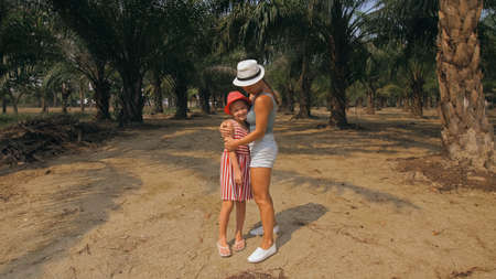 Mom and daughter for a walk. Woman tourist with plait walks looking around at growing young trees with lush leaves at oil palm farm elaeis guineensisの写真素材