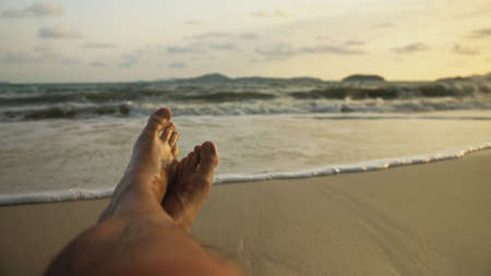 The man feet relaxed are lying on the sandy beach and washed by the water and foam of the oceanの写真素材