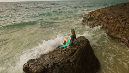 Woman sitting on rock of sea reef stone, stormy cloudy ocean. Blue swimsuit dress tunic. Concept resort coastline tourism summer holidaysの写真素材