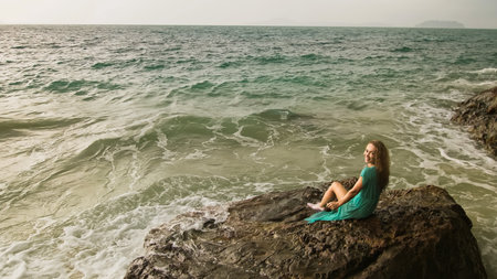 Woman sitting on rock of sea reef stone, stormy cloudy ocean. Blue swimsuit dress tunic. Concept resort coastline tourism summer holidaysの写真素材