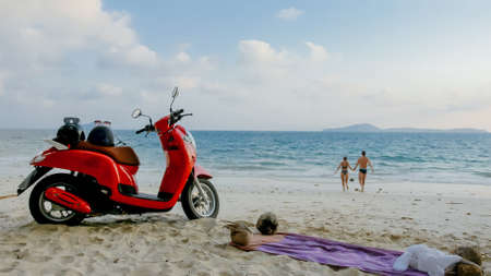 Scooter road trip. Lovely couple on red motorbike in white clothes on sand beach. People walking near the tropical palm trees, sea. Motorcycle rent.の写真素材
