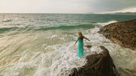 Woman walks on rock of sea reef stone, stormy cloudy ocean. Blue swimsuit dress tunic. Concept rest, tropical resort coastline tourism summer holidaysの写真素材