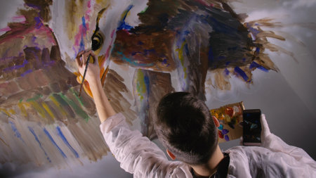 Artist designer draws an eagle on the wall. Craftsman decorator paints a picture with acrylic oil color looking at the sketch in the phone.の写真素材