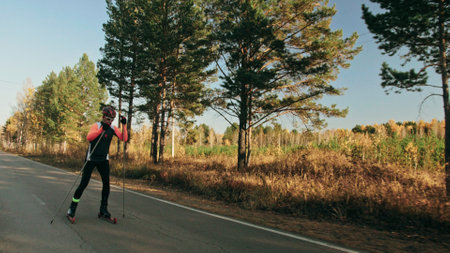 Training an athlete on the roller skaters. Biathlon ride on the roller skis with ski poles, in the helmet. Autumn workout. Roller sport. Shooting an athlete in motion. Moving platform stabilizer.の写真素材