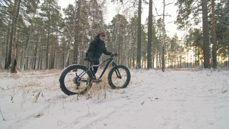 Professional extreme sportsman biker stand a fat bike in outdoor. Cyclist recline in the winter snow forest. Man walk with mountain bicycle with big tire in helmet and glasses. Slow motion in 60fps.の写真素材