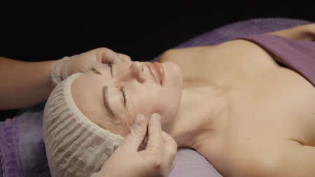 Woman receiving anti-ageing facial massage in spa salon relax. Wellness body skin care face beauty treatment. Black backgroundの写真素材