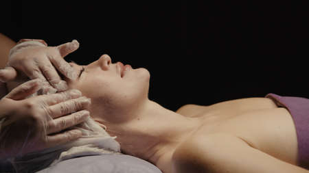 Woman receiving anti-ageing facial massage in spa salon relax. Wellness body skin care face beauty treatment. Black backgroundの写真素材