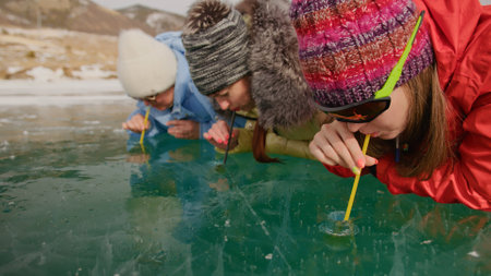 Siberia tradition cocktail Kiss of Baikal is entertainment for tourist on Lake Baikal.Three woman drink alcohol liquid without straw into makeshift glasses in hole frozen ice.Fun vacation trip travel.の写真素材